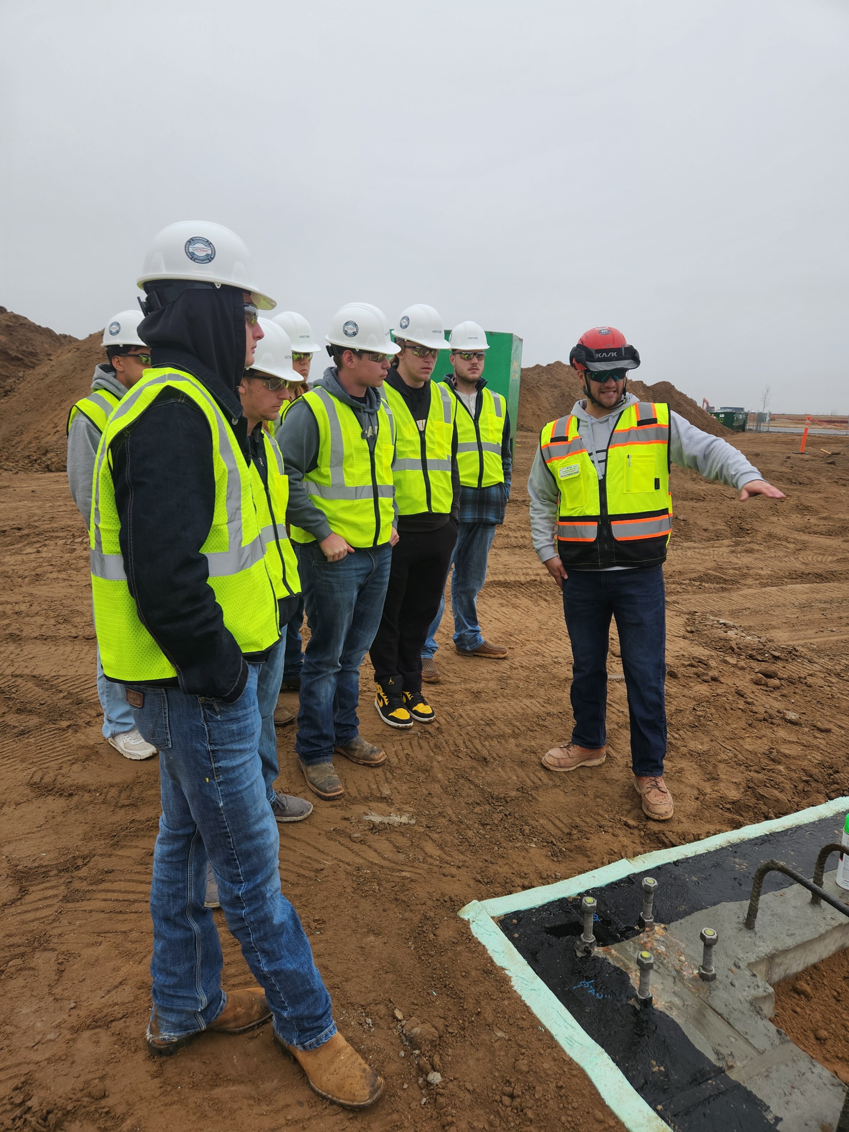 A group of students in hard hats and safety vests are on a construction site, listening to an instructor point and explain something near a concrete foundation.